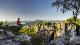 Germany, Saxony, Saxon-Switzerland National Park - A man admiring the stunning view from Malerweg towards Bastei-Felsen and Bastei bridge with Wehlnadel rock in the foreground and iconic Lilienstein, Gohrisch and Papststein peaks in the background.
